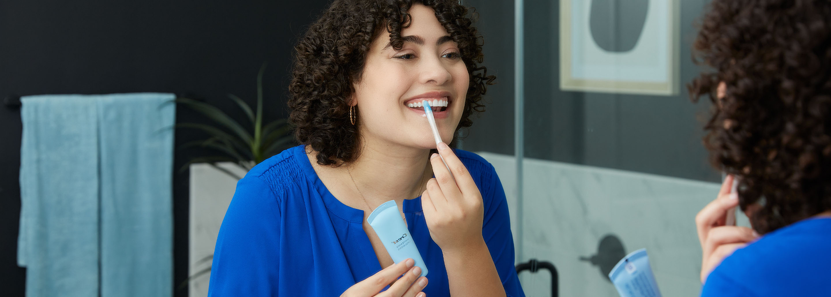 Curly-haired woman smiles while she applies Crest Whitening Emulsions with an applicator wand.