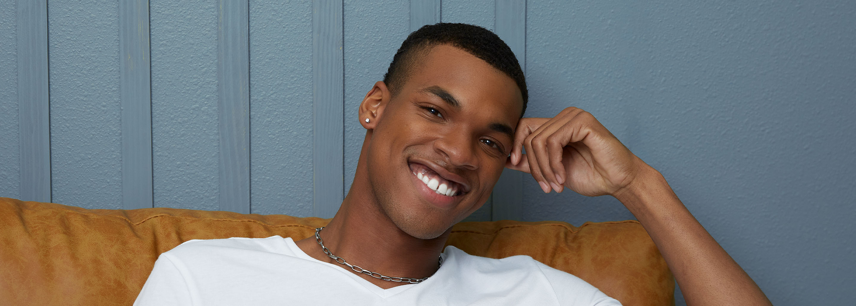 A black man smiles bright while touching his temple with one hand.