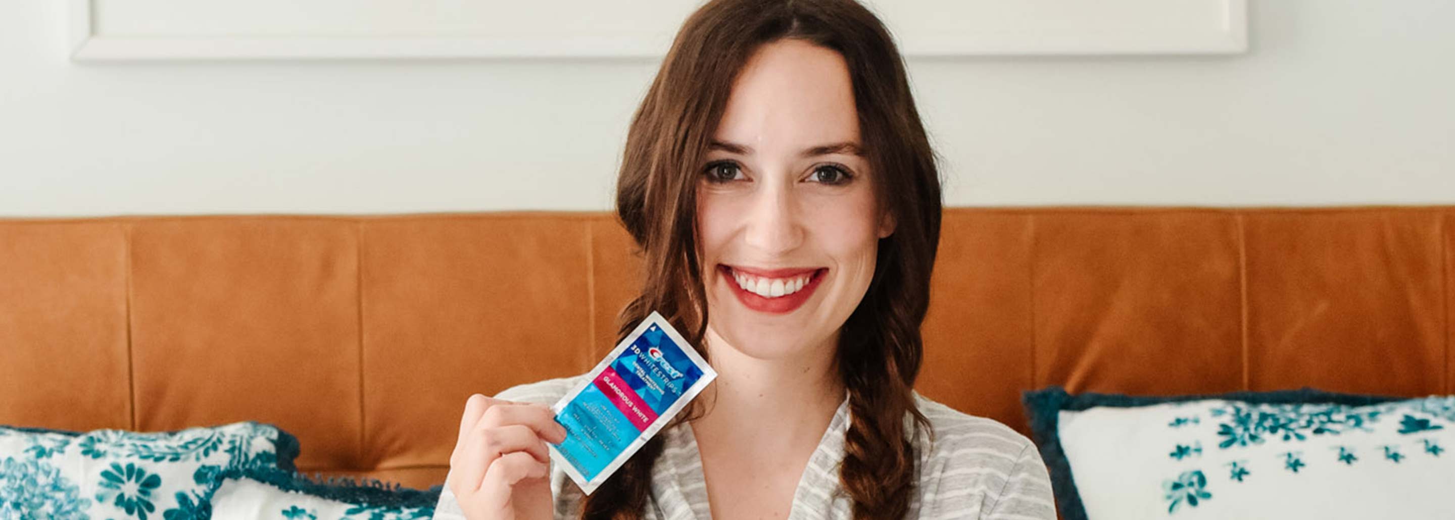 Woman with pigtail braids smiles bright as she holds a Crest 3DWhitestrips Glamorous White strip.